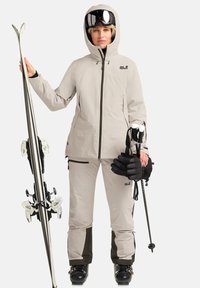 Équipement de ski comprenant une veste gris clair et un pantalon avec des accents noirs, équipé de bâtons de ski, de gants et de skis, avec une capuche et des fermetures éclair.