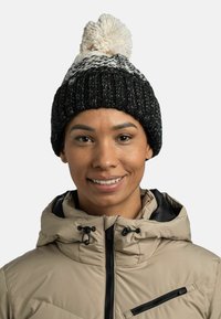 Gorro de punto negro con pompon blanco, con un patrón texturizado. Se lleva con una chaqueta beige, que muestra un cuello y detalle de cremallera.