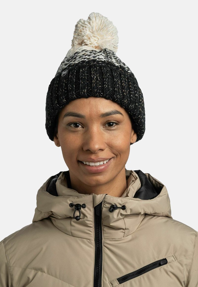 Gorro de punto negro con pompon blanco, con un patrón texturizado. Se lleva con una chaqueta beige, que muestra un cuello y detalle de cremallera.