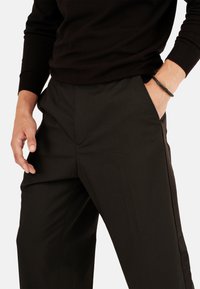 Hombre con un suéter negro y pantalones negros, con las manos descansando en los bolsillos o cerca de ellos, y lleva una pulsera trenzada negra en la muñeca derecha.