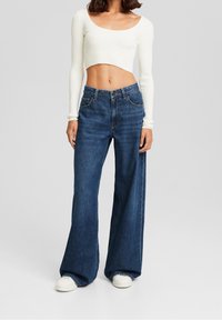 Donna che indossa un top bianco a maniche lunghe e cropped, jeans blu a vita alta a gamba larga e sneakers bianche su uno sfondo semplice.