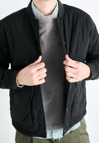 Zwarte gewatteerde jas met drukknoopsluitingen, gedragen over een grijs sweatshirt en een gestreept shirt. Heeft ribgebreide manchetten, lichtgewicht stof en een casual fit.