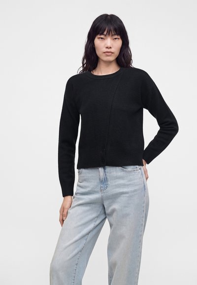 Femme aux longs cheveux foncés portant un pull noir à manches longues et un jean taille haute bleu clair, posant avec une main sur la hanche.