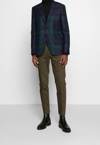 De blazer heeft een donker groene en marineblauwe ruitpatroon, een enkele knoop aan de voorkant en reverskraag. Gepaard met olijfgroene, tapered broek en zwarte enkellaarzen.