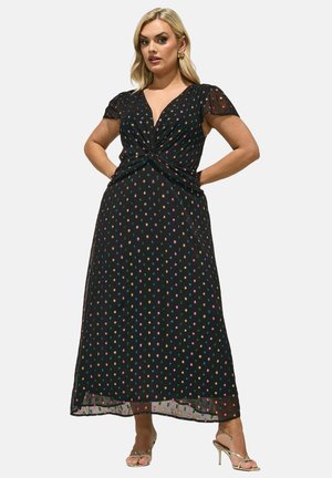 Blonde Frau trägt ein schwarzes Midikleid mit bunten Punkten, V-Ausschnitt, kurzen Puffärmeln und goldenen, Riemchen-Absatzsandalen.