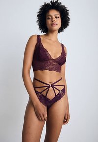 Viininpunainen pitsibralette, jossa on syvä V-aukko ja kukkakuosi, yhdistetty vastaaviin pitsialushousuihin, joissa on hihnakiinnitykset ja keskellä koriste.