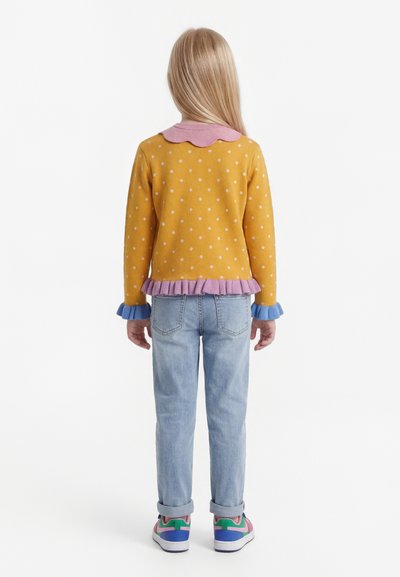 Laps, kellel on pikad blondid juuksed, seljas kollase täpilise mustriga kampsun lillade ja siniste volditud detailidega, helesinised teksad ja värvikad tossud.