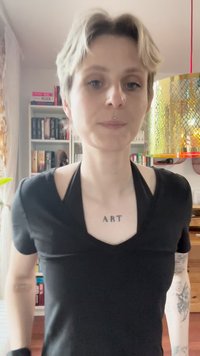 Camisa negra de cuello en V con un diseño ajustado. El tejido parece suave. Tatuaje notable "ART" en el pecho. El fondo presenta estanterías de libros.
