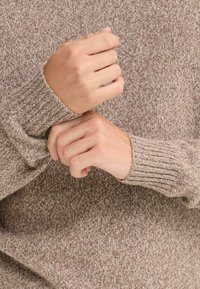 Rjav pleten jumper z teksturiranim, rebrastim manšetom. Tkanina prikazuje mešanico svetlih in temnih vlaken, kar povečuje njen globinski videz.