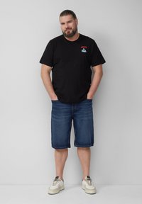 Schwarzes Baumwoll-T-Shirt mit rotem und blauem "JAWS"-Logo, kombiniert mit blauen Denim-Shorts. Modell trägt weiße Schuhe und steht lässig mit den Händen in den Taschen.