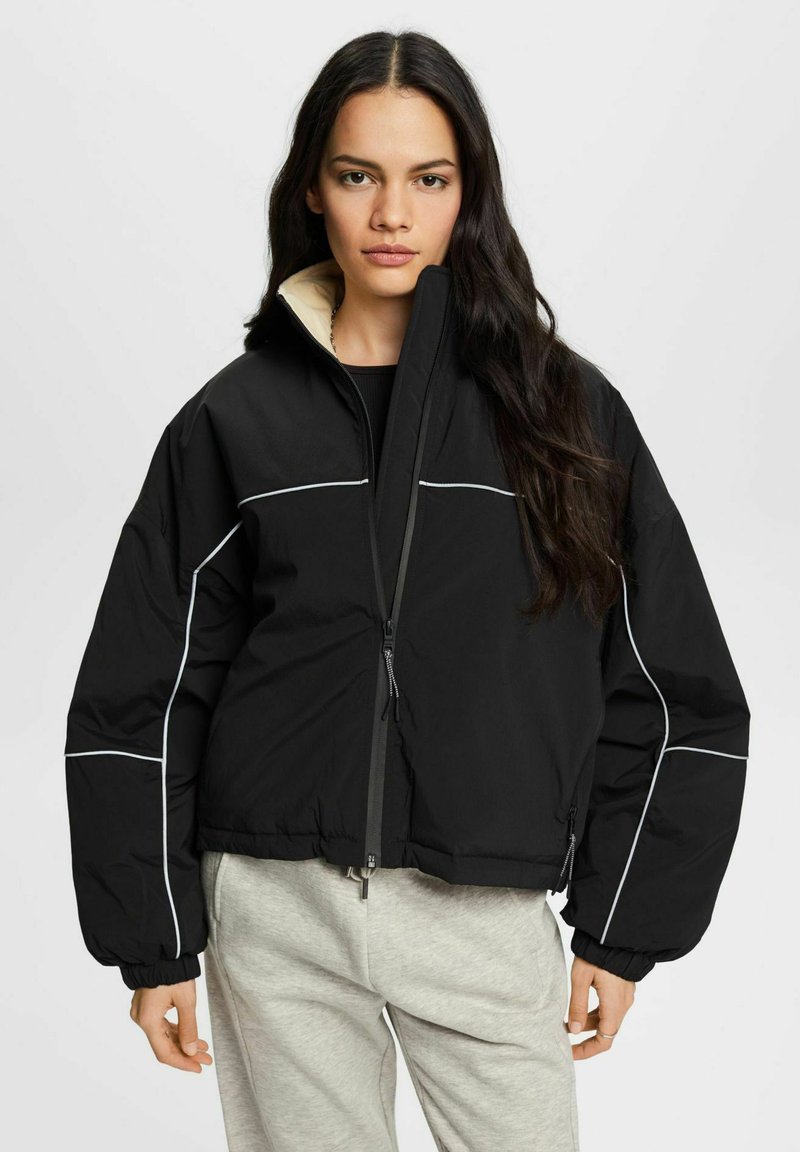 Giacca imbottita nera con colletto alto, zip frontale e dettagli in piping bianco. Maniche voluminose e design corto; foderato internamente in crema.