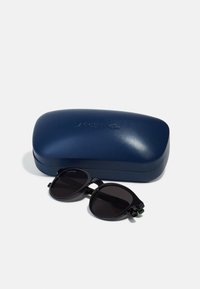 Ochelari de soare rotunzi negri cu lentile întunecate, având un mic accent verde, alături de un etui rigid bleumarin, cu înscrisul "Lacoste" embosat.