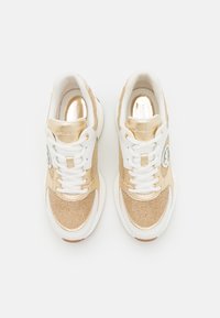 Tenisky s metalickým zlatým svrškem a texturovanými látkovými detaily, bílými akcenty, kulatou špičkou a plochými tkaničkami. Logo v blízkosti oblasti paty.