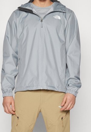 Mężczyzna ubrany w jasnoszarą kurtkę przeciwdeszczową na zamek i beżowe spodnie, oba oznaczone marką The North Face.