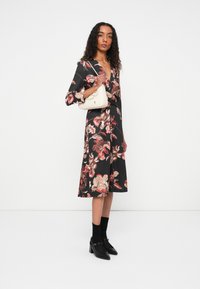 Langarm zwarte jurk met grote bloemenprint in tinten rood en beige, taille gezet, gecombineerd met een witte handtas en zwarte hoge hakken.