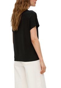 Zwart kortemouwen top met een relaxte pasvorm, gemaakt van een gladde stof. Het model draagt het met witte wijde broek. Eenvoudig en veelzijdig ontwerp.
