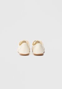 Čevlji kremne barve z gladkim usnjenim zgornjim delom, mehko tekstilno podlogo, zaobljeno obliko in svetlo rjavim gumijastim podplatom. Preprost in minimalističen dizajn.