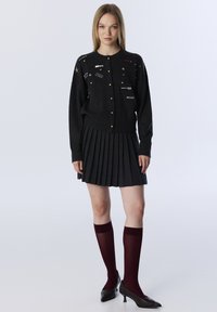 Vrouw in een zwarte knopen cardigan met patches, een geplooide donkere rok, bordeauxrode knie-sokken en zwarte hakken, staand tegen een witte achtergrond.