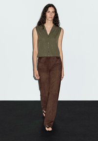 Top senza maniche verde oliva con scollo a V e dettaglio dei bottoni, abbinato a pantaloni dritti in suede marrone. Texture liscia, design minimale.