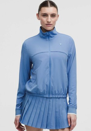 Kobieta w jasie na suwak w jasnoniebieskim kolorze i pasującej plisowanej spódnicy na jednolitym tle, zwrócona przodem z neutralnym wyrazem twarzy.