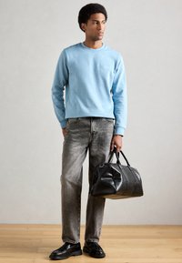 Sweatshirt azul claro de gola redonda com um pequeno logotipo, combinado com jeans cinzentos. Uma mala de viagem preta texturizada e sapatos pretos completam o visual.