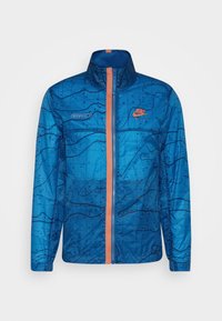 Kevyt sininen Nike-takki, jossa on abstrakti musta kuvio, oranssi vetoketju ja yksityiskohtia. Takissa on korkea kaulus ja pitkät hihat.
