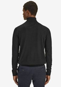 Mustat neulepusero korkealla kauluksella, pitkillä hihoilla ja ribbauslahkeilla. Sujuva tekstuuri ja slim-fit muotoilu. Pukeutunut tummansinihin housuihin.