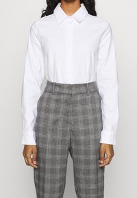 Camicia bianca a maniche lunghe con colletto abbinata a pantaloni a vita alta grigio a quadri, caratterizzati da una sottile trama e un motivo a check.
