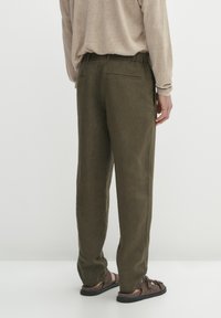 Pantaloni in lino verde oliva con vestibilità rilassata, dotati di tasche posteriori e cintura elastica, indossati con sandali marroni.
