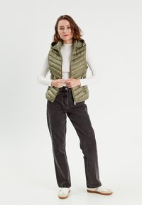 Gilet rembourré vert olive avec capuche, fermeture éclair et poches latérales, porté par-dessus une chemise à manches longues blanche et un jean noir, texture brillante.