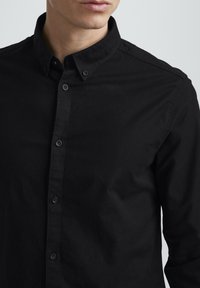Black button-up shirt made of cotton, featuring a collar, long sleeves, and a smooth texture. Includes buttons along the front.