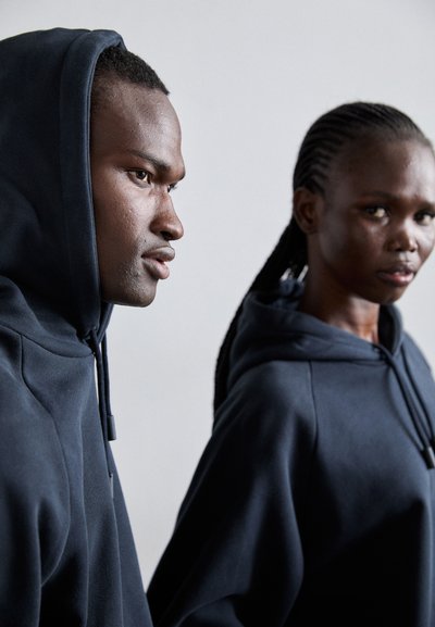 Deux personnes portant des sweat-shirts à capuche bleu foncé, avec un tissu texturé et des accents de cordon, sur un fond neutre.