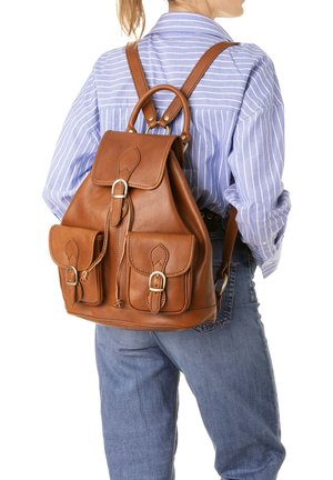 Vrouw met een blauw gestreept shirt en jeans, met een bruine leren rugzak met voorvakjes met gespen en een bovenste handvat.