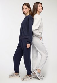 Due modelli stanno schiena contro schiena, uno indossa una felpa blu navy e pantaloni, mentre l'altro è vestito con un completo grigio chiaro, entrambi abbinati a sneaker.