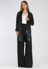 Czarny, cekinowy długi kardigan noszony na białej bluzce i szerokich czarnych spodniach. Kardigan ma luźny krój, długie rękawy i nie posiada zapięć.