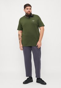 Olijfgroene T-shirt met korte mouwen en een ronde halslijn, voorzien van een klein logo. Combineert met losse grijze broek en zwarte sneakers. Sjaal om de hals geknoopt.