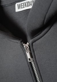 Sweat à capuche noir zippé avec une texture lisse, doté d'une fermeture éclair argentée et d'une étiquette "WEEKDAY" à l'intérieur du col.