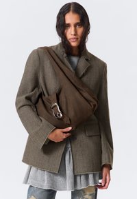 Bolso de hombro de lona marrón con una hebilla plateada, exhibido contra un modelo que lleva un suéter gris texturizado y una chaqueta de espiga, de pie contra un fondo blanco.