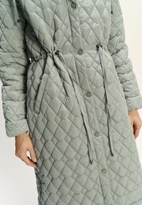 Gepolsterter, hellgrauer Mantel mit verstellbarem Kordelzug in der Taille, Druckknöpfen und langen Ärmeln mit strukturiertem Rautenmuster.
