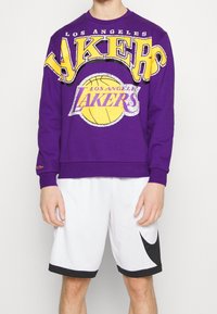 Sweat-shirt violet avec un grand texte jaune "LAKERS" et un graphisme de ballon de basket. Associé à un short blanc, mettant en valeur un design sportif.