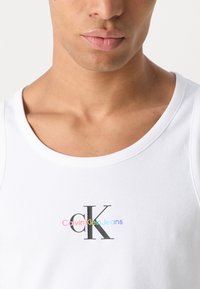 Bijela rebrasta majica na bretelama s okruglim izrezom. Na prednjoj strani nalazi se crni "CK" logo iznad šarenog teksta "Calvin Klein Jeans".