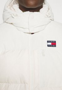 Gros plan d'une personne portant une veste matelassée blanche avec un col montant et le logo Tommy Jeans sur la poitrine.