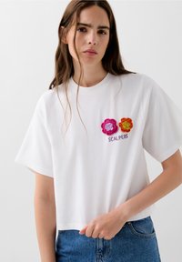 Maglietta corta bianca in cotone, decorata con fiori ricamati rosa e arancioni, e la scritta "SCALPERS" sotto il design. Maniche corte.