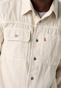 Koszulka w kremowym kolorze, wykonana z bawełny, o teksturowanym wykończeniu. Posiada dwie kieszenie na piersi, zapięcie na guziki oraz logo marki. Srebrne guziki.
