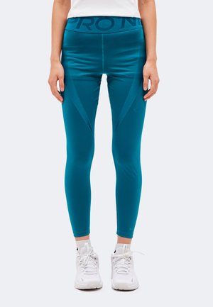 Mannequin portant un legging de sport taille haute bleu sarcelle et des baskets blanches, cadré de la taille aux pieds sur un fond clair uni.