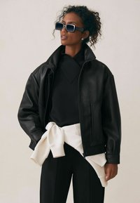 Czarna skórzana kurtka z wysokim kołnierzem, teksturowanym wykończeniem, z zapięciem na zamek i bocznymi kieszeniami. Nosi się ją na czarnym topie i białym swetrze związanym w talii.