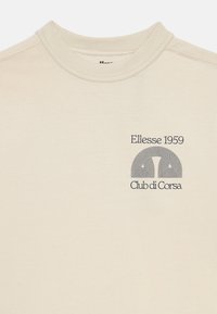 Šviesiai rudas medvilninis marškinėliai su apvaliu kaklu, kuriuose yra grafika su koncentriškomis linijomis ir tekstu "Ellesse 1959 Club di Corsa."