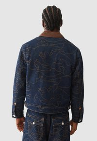 Denimjack in donkerblauw met een abstract geel lijnpatroon, voorzien van een bruine corduroy kraag en knoopdetails op de mouwen.