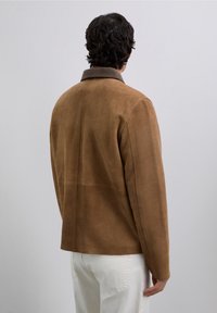 Hombre con cabello rizado oscuro, vistiendo una chaqueta de ante marrón con cuello y pantalones blancos, de pie de espaldas contra un fondo liso.