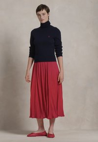 Pulover ciel cu guler înalt, asortat cu o fustă roșie lungă până la genunchi, cu un design plisat, purtată cu pantofi plat roșii asortați.
